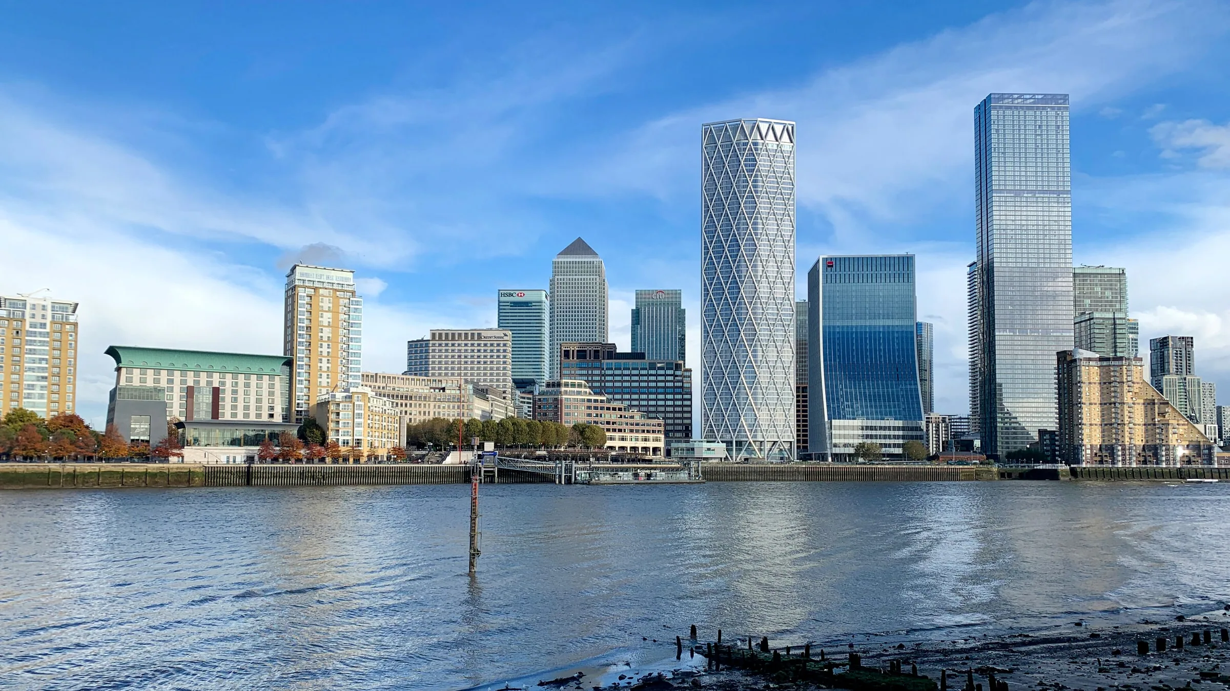 The towers of Canary Wharf across the Thames under a blue sky, seen from the exposed shore at Rotherhithe at low tide