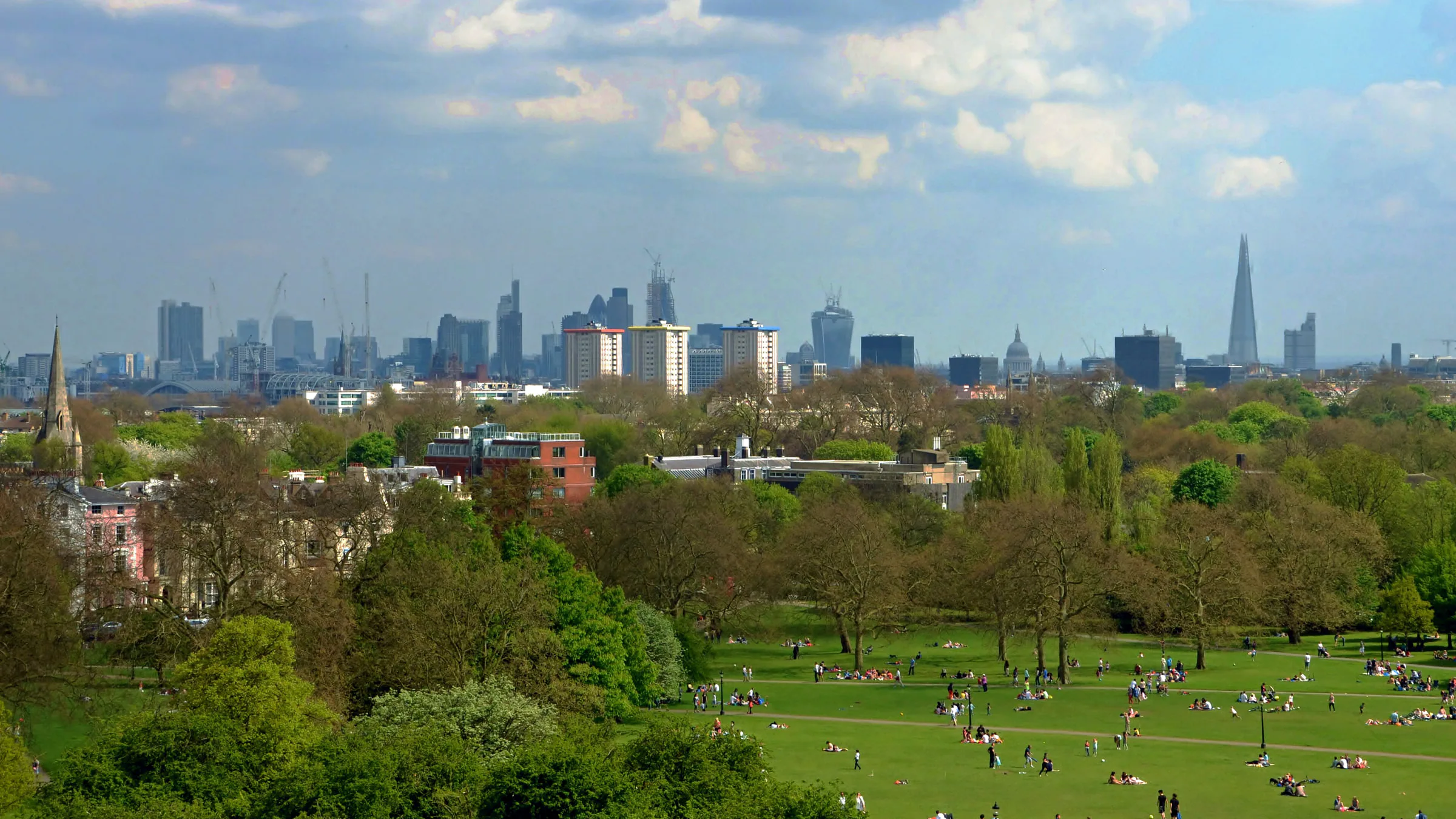 People on the grass of Primrose Hill, with trees in spring leaf and the towers of the City, St Paul's and the Shard on the skyline