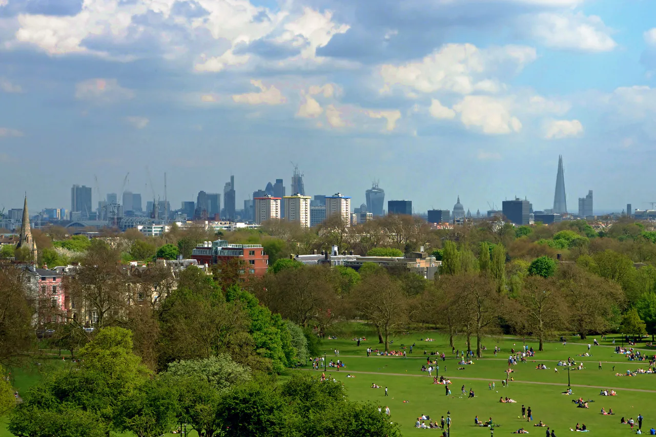 People on the grass of Primrose Hill, with trees in spring leaf and the towers of the City, St Paul's and the Shard on the skyline