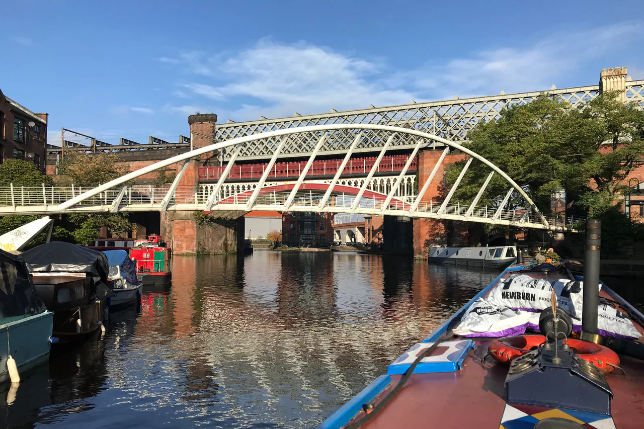 A curved white footbridge and an iron railway viaduct over the canal at Castlefield, Manchester, seen from the bow of a narrowboat