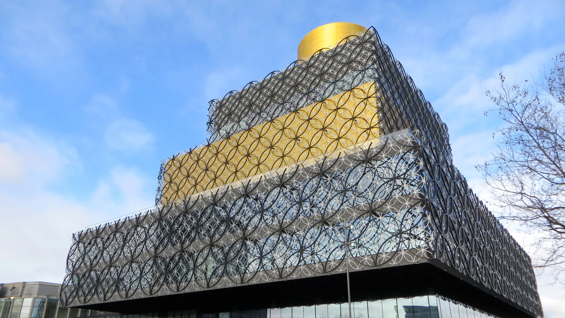 The Library of Birmingham, its stacked boxes wrapped in silver and gold metal rings under a gold drum on the roof, against a blue sky