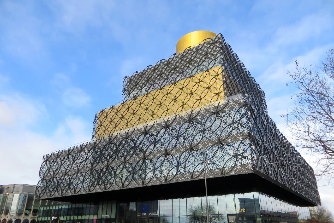 The Library of Birmingham, its stacked boxes wrapped in silver and gold metal rings under a gold drum on the roof, against a blue sky