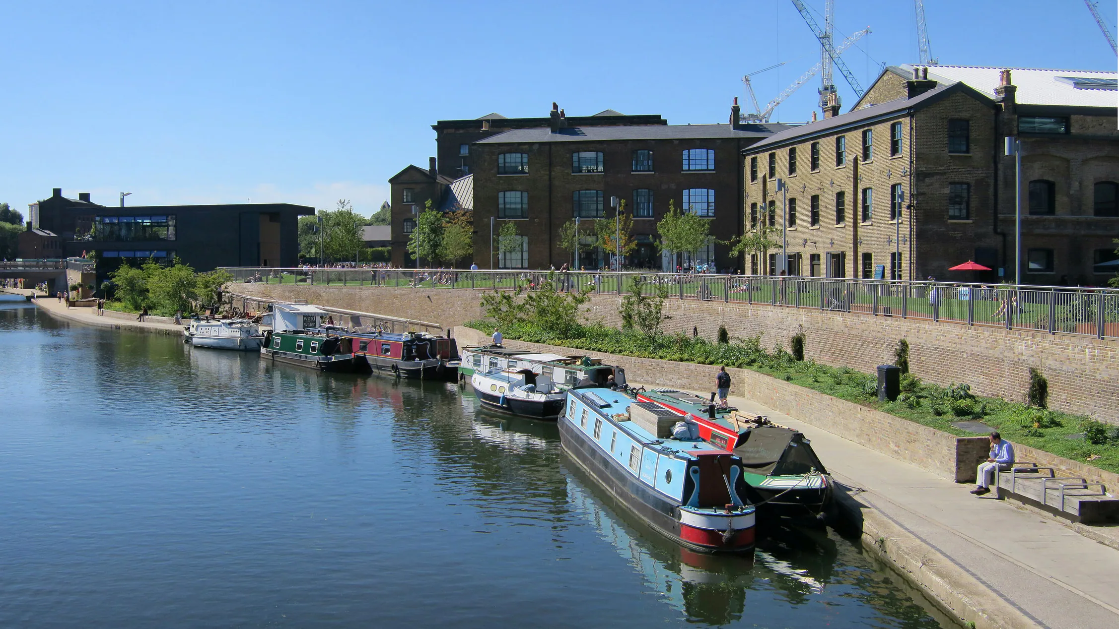 Narrowboats moored on Regent's Canal below grass steps where people sit in the sun, with brick warehouse buildings and cranes behind