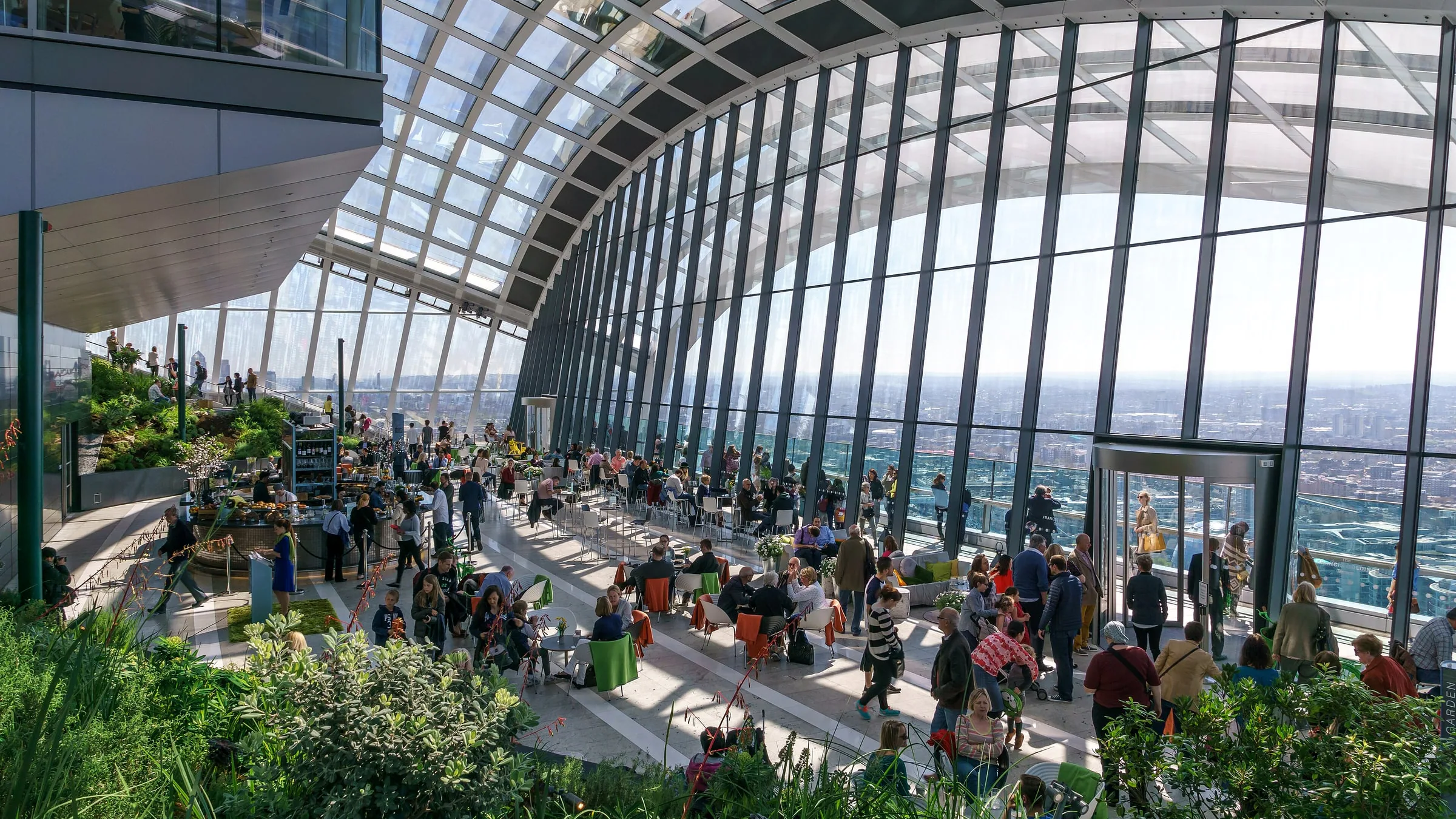 Visitors at tables and among planted terraces under the curved glass roof of the Sky Garden, with London spread out below