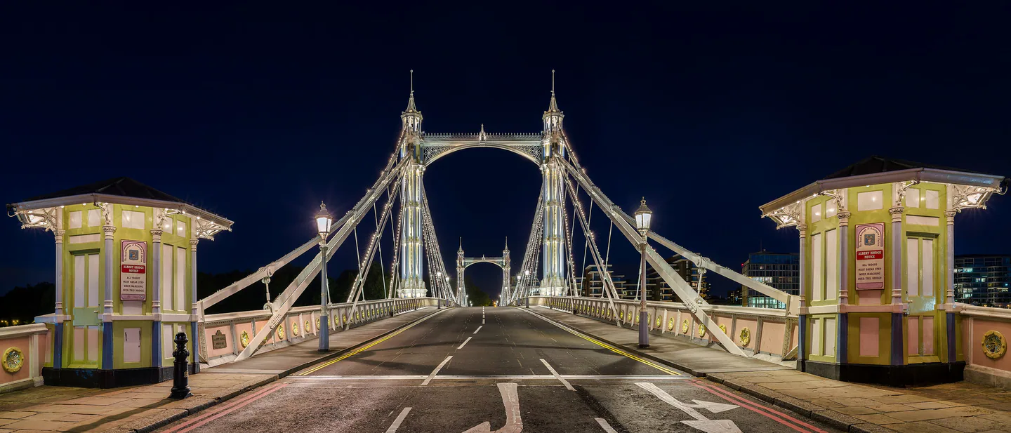 Albert Bridge lit up at night, its white towers and cables framing the empty road, with a pastel toll booth at each side
