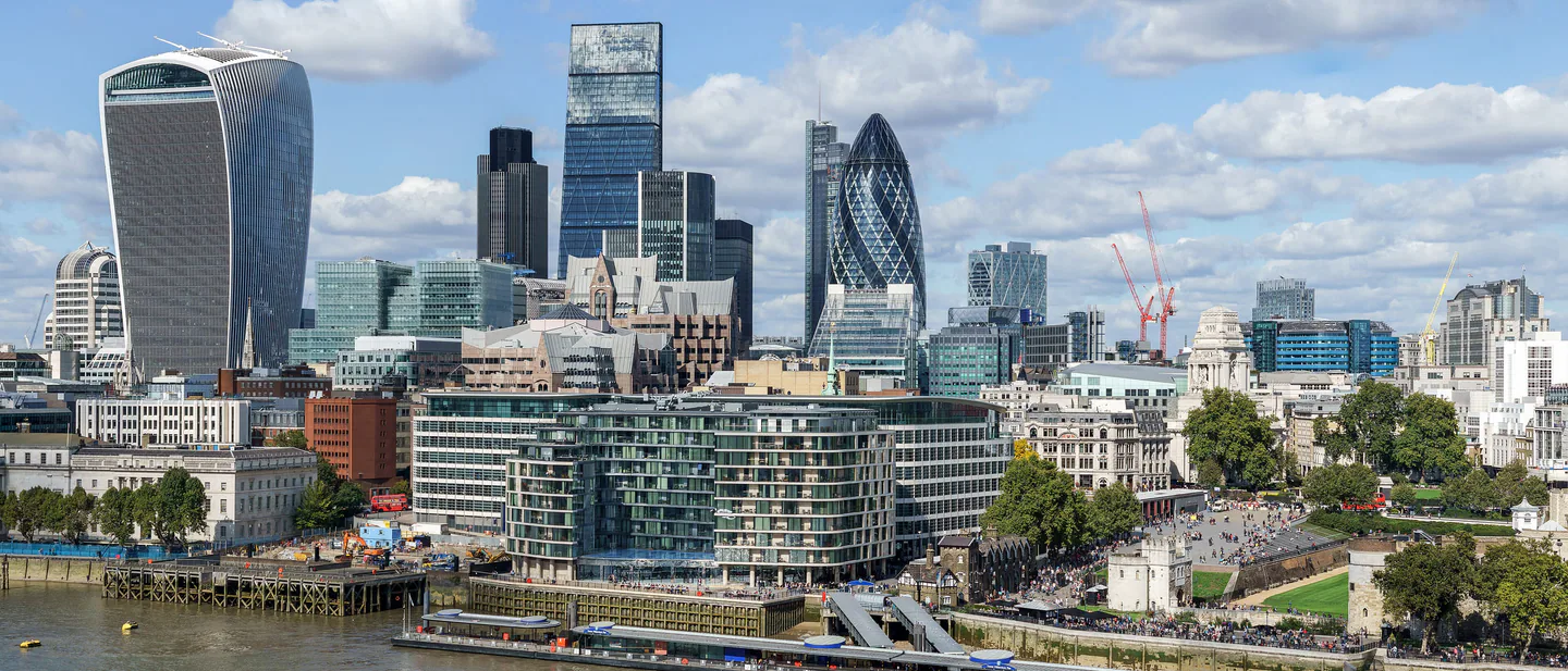 The towers of the City of London, the Walkie-Talkie and the Gherkin among them, rising above the Thames and Tower Millennium Pier under a blue sky with white clouds