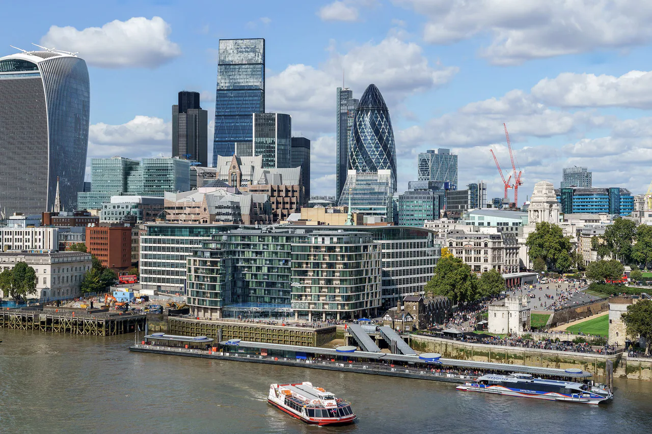 The towers of the City of London, the Walkie-Talkie and the Gherkin among them, rising above the Thames and Tower Millennium Pier under a blue sky with white clouds