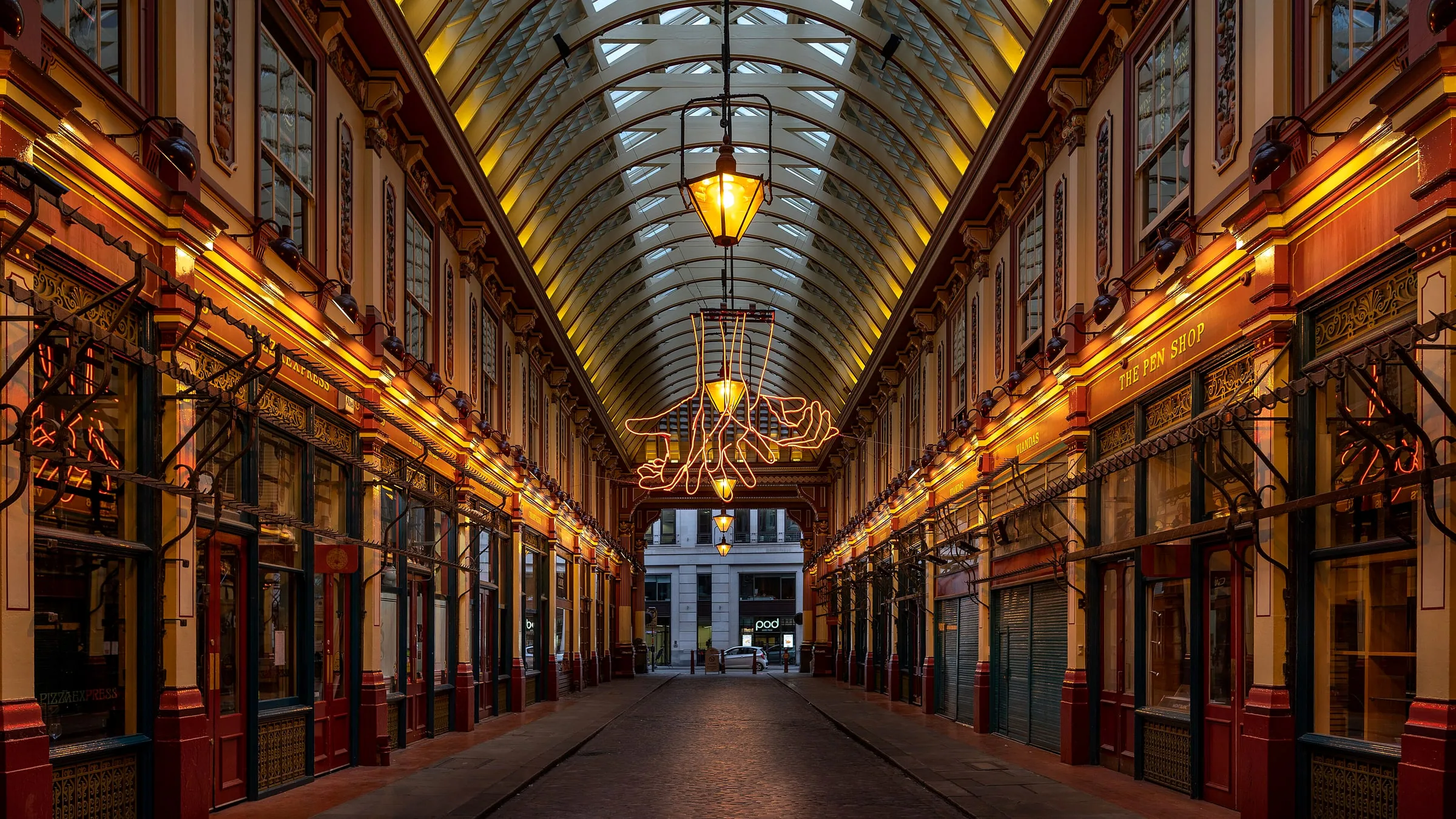 The empty cobbled walkway of Leadenhall Market under its lit glass roof, with painted shopfronts on both sides